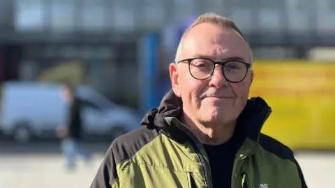 Tudor Evans wearing black-framed glasses and a black crewneck top with an olive green and black hooded jacket againsta. blurred background.