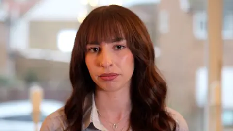 Daisy Foster looks at the camera, the background is out of focus but seems to show low rise homes through a window. Daisy has long dark hair which is wavy and has a fringe.