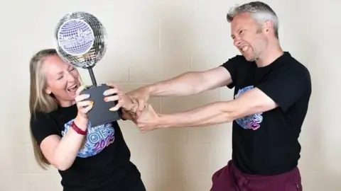 Kelly and Rob and standing next to each other wearing black t-shirts that say "dance floor heroes 2025". Kelly is holding onto a trophy shaped like a disco ball. Rob is trying to wrestle it off her