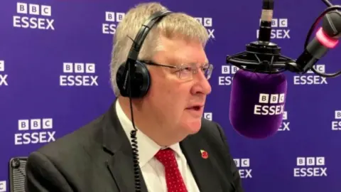 Roger Hirst  with grey hair wearing a black suit jacket, a white shirt, and a red tie.