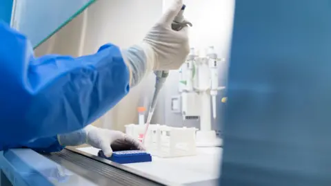 A worker in blue overall and white gloves is working in a lab. There is scientific equipment in front of them and they are holding a large syringe. It has pink liquid which is being added to a blue tray.