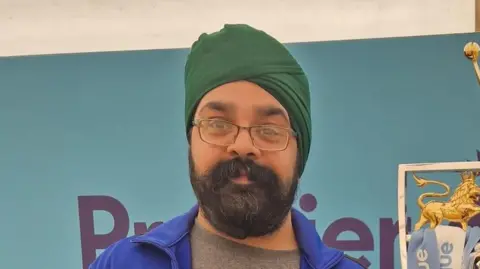 Supplied A head and shoulders image of a man. He has a green turban, glasses, and a thick beard. He is looking into the camera. 