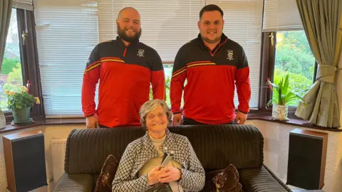 Two men in rugby uniform stand behind a sofa in a living room, and an elderly woman smiles at the camera, sitting on the sofa holding a TV remote 