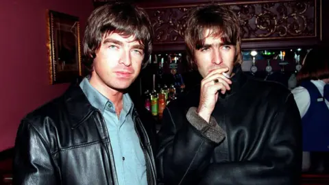 Getty Images Picture of Liam and Noel at the opening night of Steve Coogan's comedy show in the West End, London in 1995. They are stood together with a bar behind them. Liam, on the left, is wearing a blue shirt and a black leather jacket and is looking at the camera. Noel, on the right, is looking away from the camera and is holding a cigarette up to his mouth. He is wearing a black leather jacket.