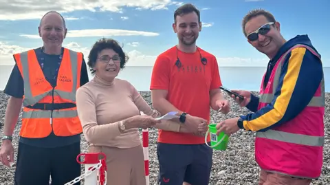 Seaton Parkrun Joan Norcombe at the end of the Seaton Parkrun with three male marshals, two of whom are wearing high-vis jackets.