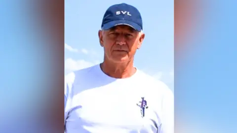 Veteran Steve Coombs wearing a navy blue cap with the logo SVL on it and a white t-shirt looking straight-faced and standing in front of blue sky with some clouds. 