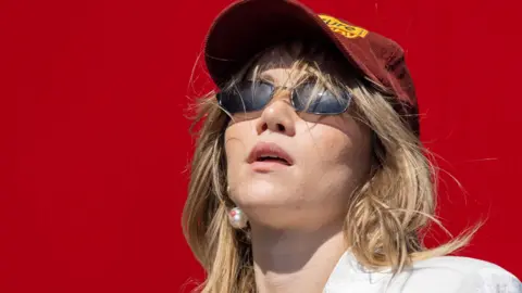 Getty Images Suki Waterhouse performing on stage in 2023. Suki is a 32-year-old woman with long blonde hair worn loose under a burgundy baseball cap. She wears small dark sunglasses, a white shirt and pearl earrings. She's pictured against a red background. 
