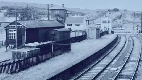 A black and white photo of a railway station. This picture has many different outbuildings surrounding the railway tracks. Next to the track is a long platform 