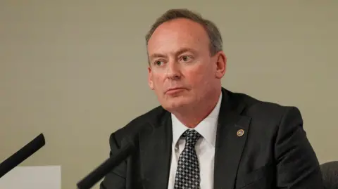 A mid-shot of Deputy Lyndon Trott. He has short dark hair, is clean shaven, and wearing a dark jacket, light shirt and tie.