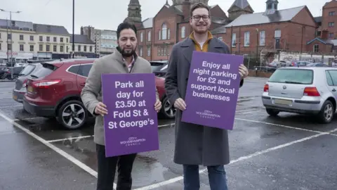 Councillor Qaiser Azeem and councillor Chris Burden stand in Fold Street car park near some cars holding purple signs advertising the new prices for shoppers and visitors. It is a grey, wet day and they have red-brick buildings and and white-painted behind them. Azeem has black hair a beard and is wearing a grey jacket. Burden has glasses on, a beard and a long grey jacket on.