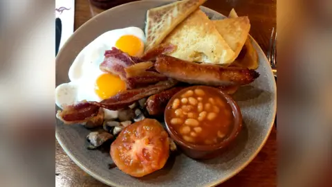 Getty Images A picture of an Ulster Fry - including beans, sausages, two fried eggs, mushrooms, potato bread, soda farls and a small bowl of baked beans on a grey stone plate.