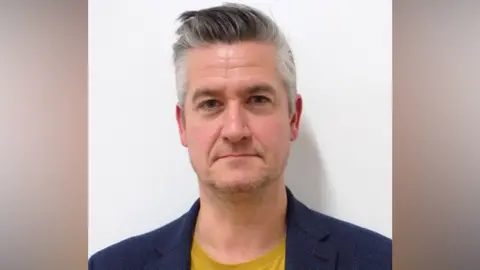 Ryan Hook stood against a white wall. He has white and grey hair styled in a quiff, wears a mustard T-shirt underneath a blue blazer.
