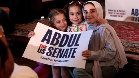 Getty Images Three young girls, two with pink hair clips and one with a beige headscarf, smile and hold a sign saying "Abdul for US Senate". 
