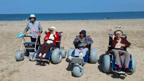 Fleetwood Beach Wheelchairs Four older people are using beach wheelchairs. They are lined up on the sand, with the sea behind them. A man on the left is using one that a support frame, while three people next to him are sitting in beach wheelchairs. They are all smiling.