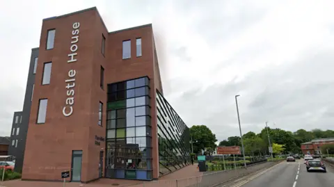 A red brick building next to a road with two lanes. There is a black sign written vertically down the tallest side of the building that reads "Castle House". The right-hand side of the building nearest to the road is made up of black glass windows. 