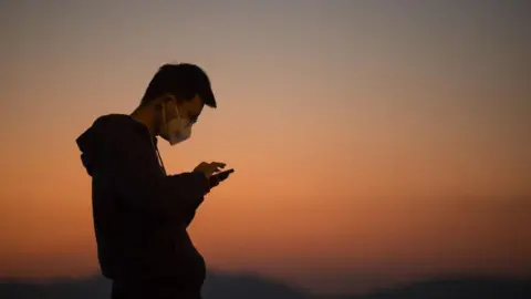 A man in a hooded sweater and a face mask uses the touchscreen of his phone in front of a sunset sky