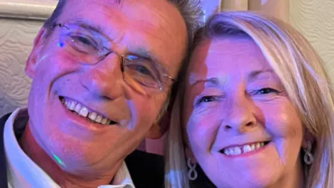 Stuart Raynor A close-up selfie of a happy couple. The man on the left had glasses without frames and wears a white shirt. The lady on the right has tidy blond hair and jewelled earrings shaped like infinity symbols. They are leaning their heads together.