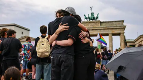 Omer Messinger/Getty Images Three people hug each other with a rainbow LGBT flag visible in the background and the Brandenburg Gate behind