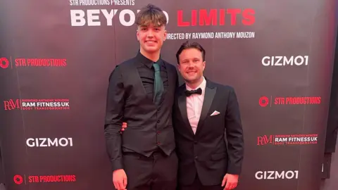 Martin Edwards Photography A tall young man wearing a black suit and a dark green tie and a slightly older man wearing a tuxedo. They're standing with their arms around each other and smiling at the camera, with a banner for their film behind them.