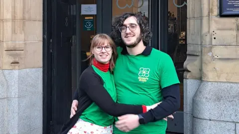 Kitti Szep A couple with their arms around each other smile at the camera. They are standing in front of a door which leads to their shop. Both are in green t-shirts which have the name written on them, and black long sleeved tops. 
