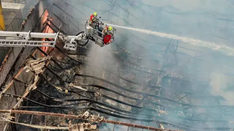 Stephen Huntley/BBC A number of firefighters on an aerial platform using a hose to extinguish a fire at a bathroom showroom on Selbourne Road in Luton on Saturday. The picture has been taken by a drone from above and there is extensive damage to the building which has collapsed in on itself.
