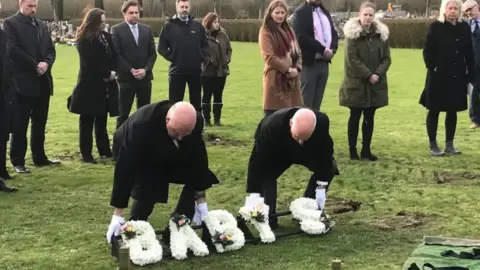 Luke Deal/BBC Mourners gather at a graveyard as two men lay a flower wreath which reads Baby S