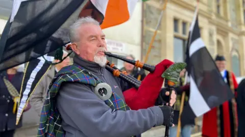 A man with white hair and a white beard playing the bagpipes, while people stand behind him holding Cornish black and white flags on wooden poles.
