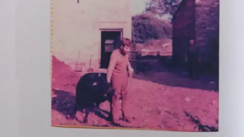 Jonathan Charlesworth A photo showing a man, holding an animal in his right hand, in front of a stone building.