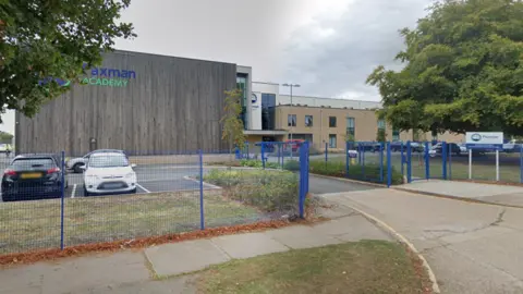 Google The front of Paxman Academy in Colchester. The school is surrounded by blue fences in front of a car park. The main school building has a wood-effect facade with the school's logo in the top left corner.