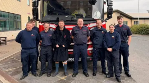 A gathering of firefighters in dark blue shirts and trousers in front of a red fire engine in a car park with a man in a black dressing gown standing amongst them