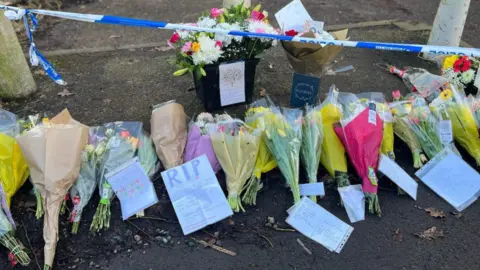 At least 16 bunches of flowers on the ground. Police tape is higher up on the photo. The tape is stretched between concrete bollards.