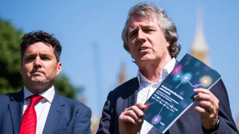 PA Huw Merriman (left) and Steve Rotheram (right). They are on College Green at Westminster, so the House of Commons is in the background out of focus. Steve Rotheram is holding a booklet about rail growth called "Connecting the North West to drive national prosperity".