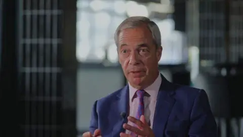 BBC Nigel Farage being interviewed by the BBC. He is looking intently, and gesturing with his hands. He is wearing a blue suit, patterned tie and pink shirt. 