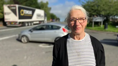 Anne Ridley standing next to the A66 with busy traffic behind her. She has white hair tied back. She wears glasses and golden hoop earrings, she has a black and white striped t-shirt with a black cardigan over it.