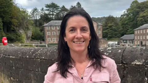 Eilis Scott wearing a pink blouse looking straight at camera in front of walls and buildings of Cromford Mills World Heritage site