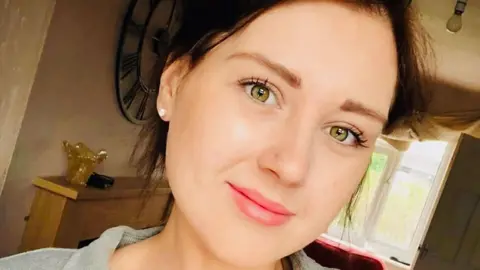Emily Rhodes A young woman indoors smiling softly at the camera, wearing a light grey shirt over a white top.