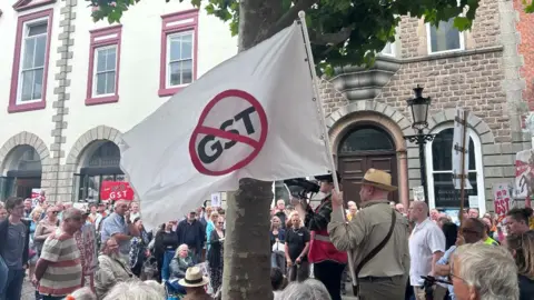 A number of people gather in a town square. In the foreground is a white flag containing a red circle with a red line through it, like a traffic sign. The letters "GST" are scored through by the line.