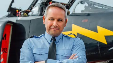 RAF Cosford A man with short grey hair, wearing a pale blue military shirt with a black tie, stands smiling at the camera with his arms cross in front of his chest. Behind him is a black military looking plane with yellow lighting stripe across it.