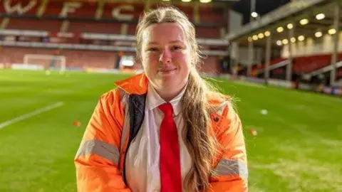 Chloe Lycett stood on the pitch at Walsall Football Club wearing a high-vis jacket and red tie. She has her hair tied back and is looking at the camera smiling. You can see the goal and stands behind her. 