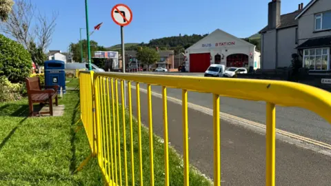 Yellow barriers line a grassy area on the left with a wooden bench and blue waste bin. There is a pavement and road on the right. There is a road sign indicating no left turn ahead and to the right further away is a large building with a pitched roof and red letters on the front reading: bus station. It is a sunny day and the area is built up and there are cars parked and using the road.