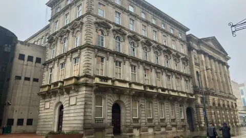 LDRS A large, historic stone building on a city street corner. The architecture is grand and ornate, with multiple floors featuring tall, arched windows framed by decorative stonework. The façade has strong classical influences, including carved details above the windows and prominent vertical pilasters that give the building a sense of height and solidity. The lower level is built from heavier, more textured stone blocks, and the entrances are set within tall arched doorways with dark wooden doors. Higher up, the stone becomes smoother, and each floor has evenly spaced windows that add to the building’s symmetrical appearance. The sky is overcast, creating a muted, slightly foggy atmosphere, and the pavement appears damp, suggesting recent rain. A couple of people are walking on the right side of the image, giving a sense of scale to the building. On the left, part of a more modern structure is visible.