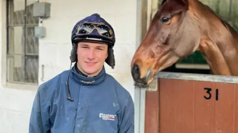 A picture of a man wearing a navy blue jacket and a blue helmet standing outside a stable with the number '31' on it. There is a brown horse inside the stable