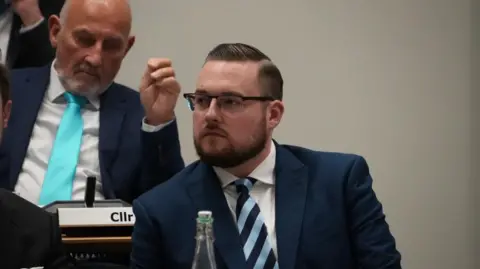 John Fairhall/BBC Michael Hadwen is sitting in a council meeting and is wearing a blue suit and white shirt with a blue and teal striped tie. 