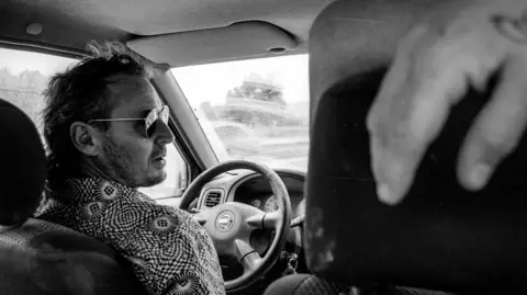 A picture of a man in black and white sat in the front seat of a car.