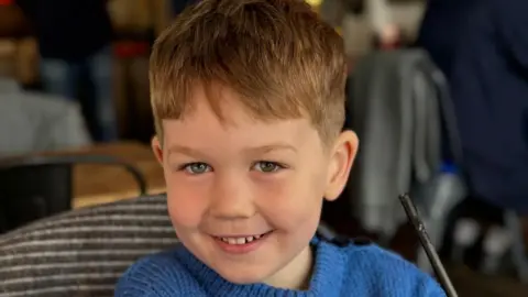 Leanne Rinne/BBC A young boy smiling with blonde hair wearing a blue jumper 