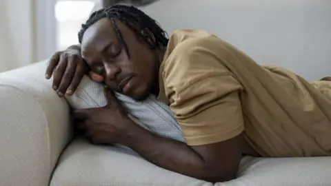 Getty Images A man sleeping on a sofa at home with a grey cat resting in the background. His relaxed posture and closed eyes suggest a peaceful nap, rest, fatigue, or quiet leisure time