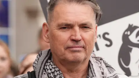 Dale looks at the camera with a serious expression. He has grey hair and wears a white and black patterned scarf. 