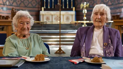 The two codebreaking ladies sitting in the church where they have been presented with an award. Kath is wearing a green top while Gwenfron is wearing a purple jacket, both have short white hair. There is cake laid on the table along with a framed a certificate and a book.