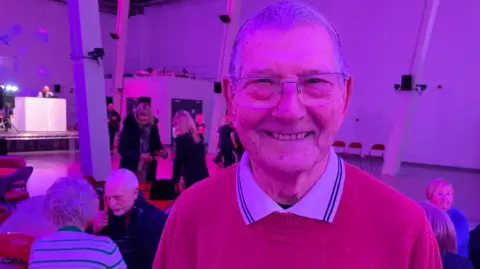 Tom Ingall/BBC A man with grey hair and glasses, in a red jumper and white polo shirt, at a hall which has pink lights and is being used as the venue for a disco for older people