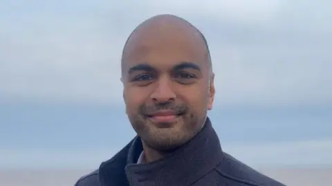 Contributed Ben Ponniah smiles at the camera. He is standing outdoors and the sky above him is cloudy and grey. He wears a dark coat. 